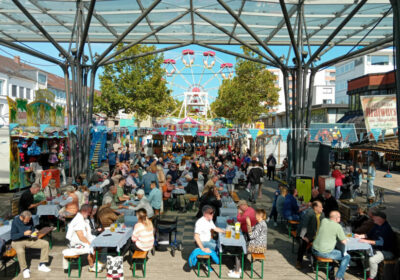 Menschen auf Bierbänken unter einem Glasdach und rundherum Oktoberfest-Stände und bayrischen Dekoration.