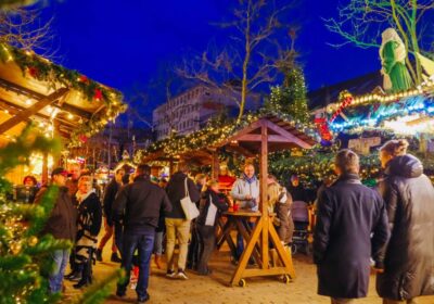 Belebter Wolfsburger Weihnachtsmarkt am Abend mit festlich beleuchteten Holzständen, dekorierten Tannenbäumen und Besuchenden, die in gemütlicher Atmosphäre zusammenstehen.