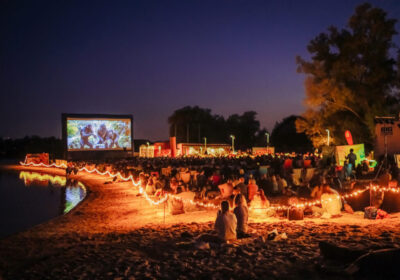 Große Leinwand auf einem Strand und Wasser daneben. Das Gelände ist stimmungsvoll beleuchtet und mehrere hundert Menschen sitzen auf Liegestühlen und gucken sich einen Film an.