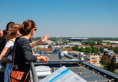 Personen genießen bei einer Führung den Ausblick über Wolfsburg von einer Dachterrasse aus und zeigen in Richtung Stadtpanorama.