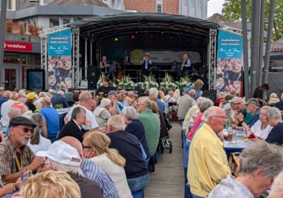 Jazzband spielt auf Bühne und davor sitzen viele Menschen auf Bänken an Tischen und unterhalten sich.