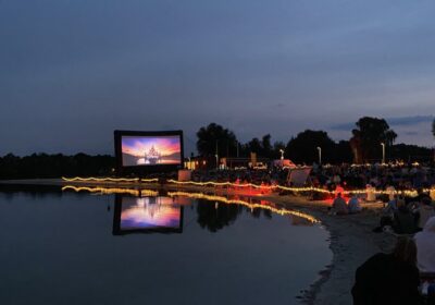 Im Hintergrund eines Ufers steht eine große Leinwand am Strand. Davor sitzen hunderte Menschen in Liegestühlen in der Abenddämmerung.