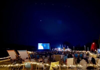 Im Hintergrund steht eine große Leinwand am Strand. Davor sitzen hunderte Menschen in Liegestühlen. Es ist dunkel.